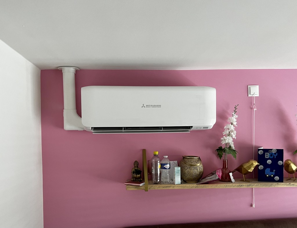 Mitsubishi Heavy wandunit binnenunit, gemonteerd aan een roze muur in een kamer. De airco heeft een modern, strak ontwerp met een luchtrooster aan de voorkant voor efficiënte luchtverdeling en temperatuurregeling.