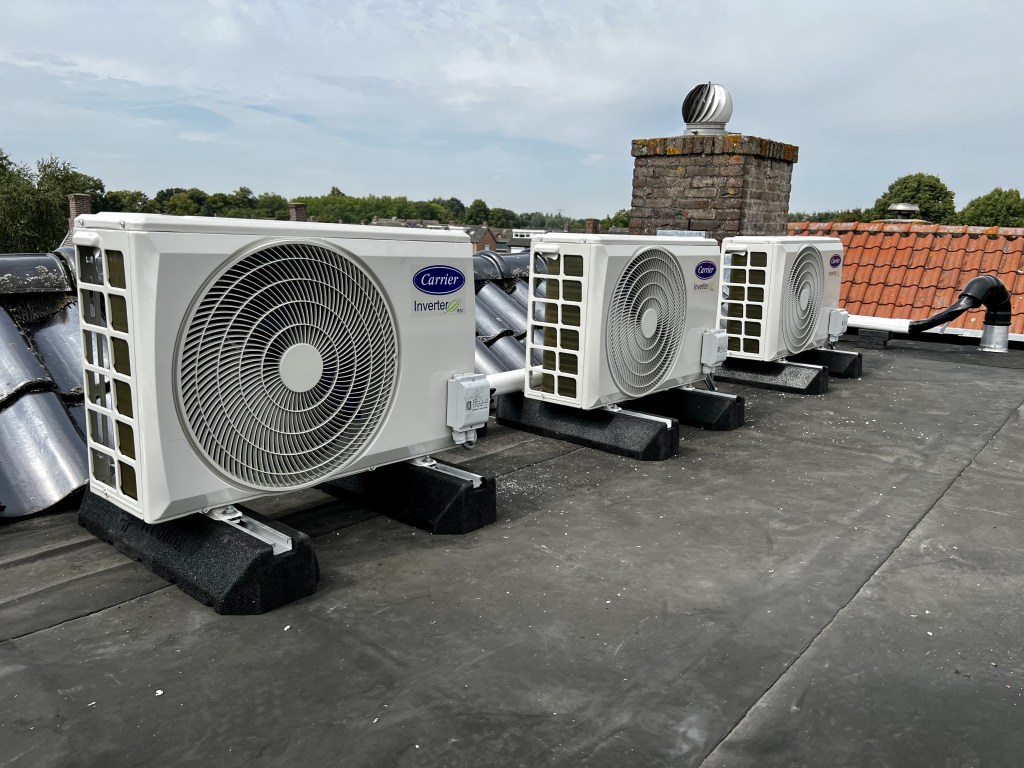 Drie Carrier buitenunits van single split airconditioningsystemen, opgesteld in een rij op een dak. De units hebben een robuust, compact ontwerp en zijn voorzien van ventilatoren voor efficiënte koeling en verwarming, geïnstalleerd voor optimale luchtcirculatie.