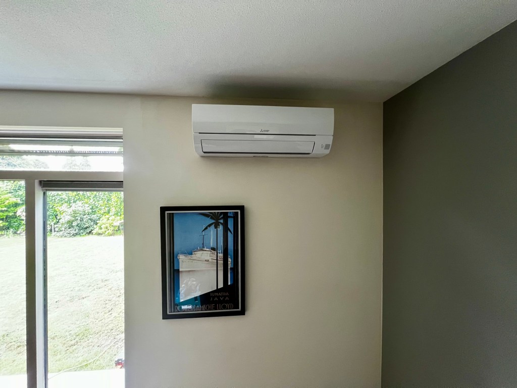 Wandunit, binnenunit, van een Mitsubishi Electric airconditioningsysteem, gemonteerd aan de muur in een kamer. Het heeft een strak, modern ontwerp met een breed luchtrooster aan de voorkant voor efficiënte luchtverdeling en temperatuurregeling.