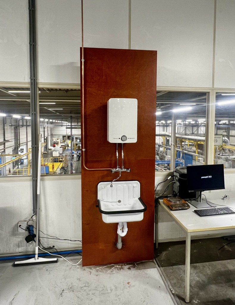 Close-up van een ruimte in een bedrijfspand waar loodgieterswerkzaamheden zijn uitgevoerd, met zicht op een boiler, uitstortgootsteen, wastafel, kraan, waterleiding, afvoer/ riolering.